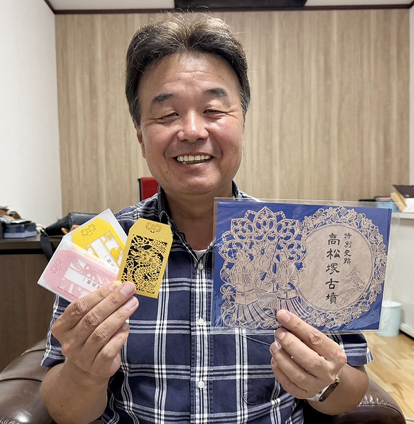 高松塚古墳の御朱印㊨と一願成就お守りを持つ浪江社長